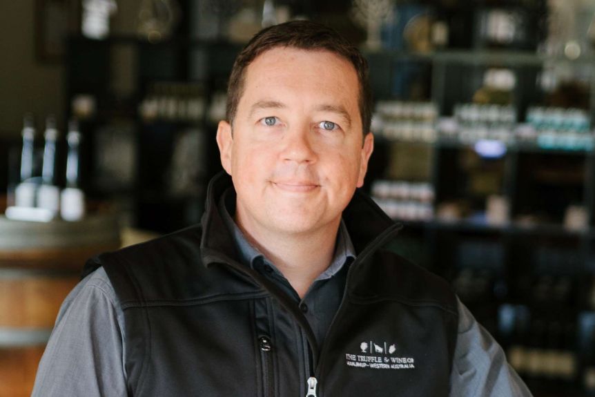 A headshot of a man with a vest on with a blurred background of products in the background