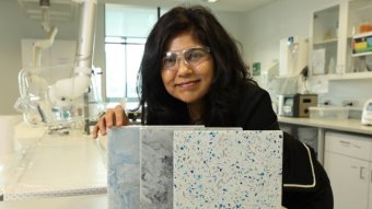 A woman wearing clear safety glasses smiles in a lab next to three marble-like tiles.