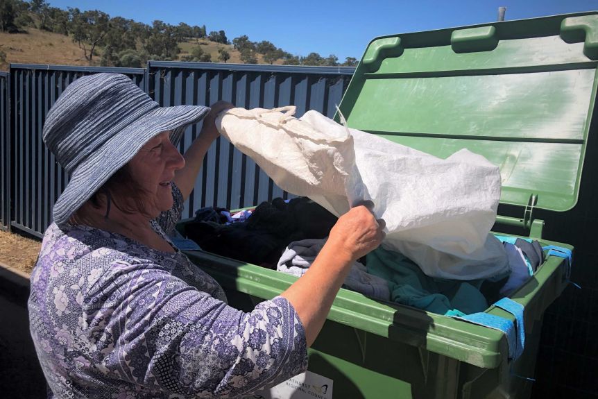 A woman in a blue hat tips clothes out of a white polyester bag into a large green bin.